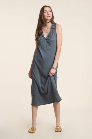 A woman in the YORK DRESS by Velvet by Graham & Spencer, a sleeveless midi dress, wears yellow sandals and stands against a plain beige background.