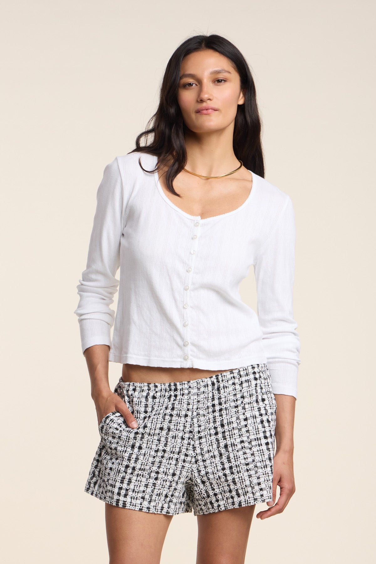 A woman stands with one hand in her pocket, wearing the ZELDA CARDIGAN by Velvet by Graham & Spencer—a white cotton cardigan with scallop cuffs—paired with black-and-white patterned shorts, set against a neutral background.