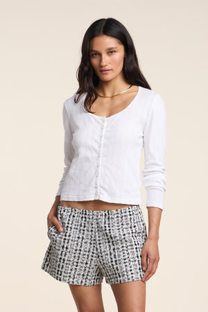 A woman wears the ZELDA CARDIGAN by Velvet by Graham & Spencer, a white long-sleeve top with scallop cuffs, paired with black-and-white patterned shorts, standing against a plain light background.