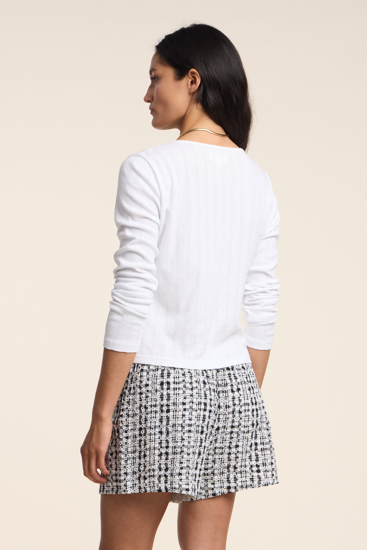 A woman stands with her back to the camera, wearing the ZELDA CARDIGAN by Velvet by Graham & Spencer— a white cotton cardigan with scallop cuffs—paired with black-and-white patterned shorts against a plain light background.-41527126950081