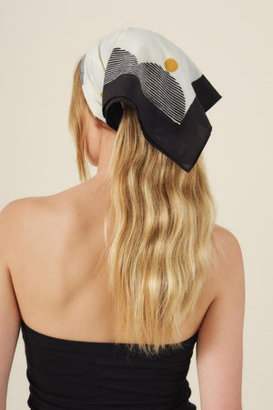 A woman with long, wavy blonde hair wears the ZURI SCARF by Velvet by Graham & Spencer over her head and a black strapless top, photographed from behind against a neutral background.