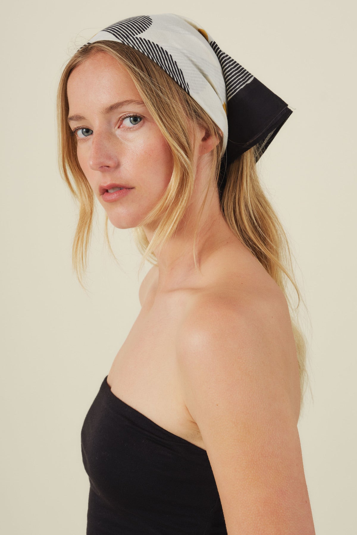 A woman with long blonde hair wears the ZURI SCARF by Velvet by Graham & Spencer as a stylish accessory with a black strapless top, looking over her shoulder against a plain background.