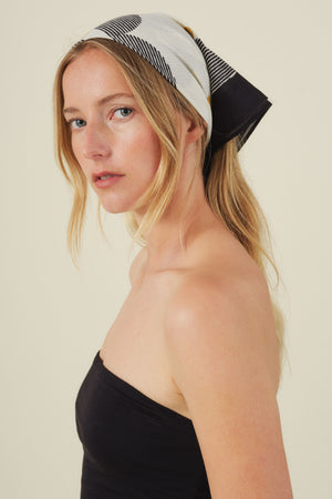 A woman with long blonde hair wears the ZURI SCARF by Velvet by Graham & Spencer as a stylish accessory with a black strapless top, looking over her shoulder against a plain background.