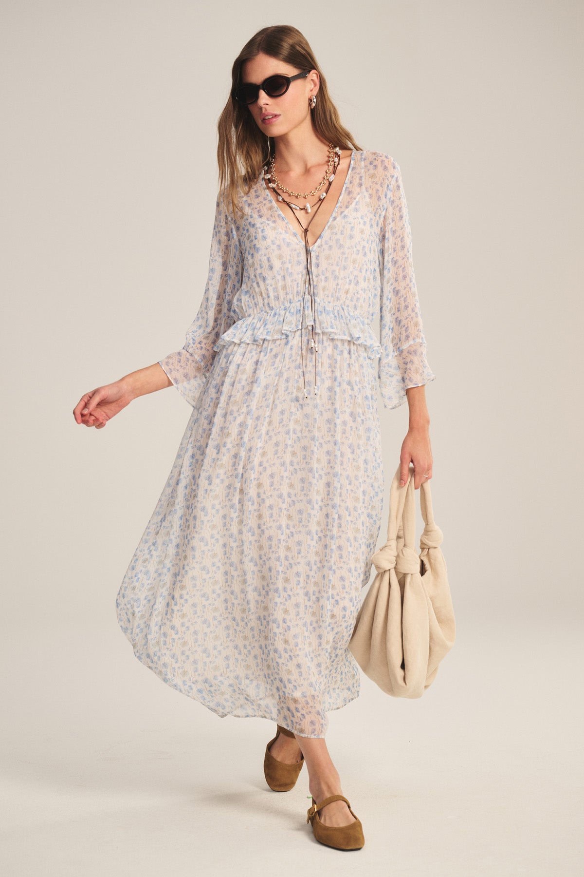 Wearing the AERIN DRESS by Velvet by Graham & Spencer, a woman in brown flats, dark sunglasses, and layered necklaces holds a beige knotted tote bag as she stands against a plain background.-40909523845313