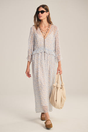 A woman wearing sunglasses and the AERIN DRESS by Velvet by Graham & Spencer with layered necklaces, brown shoes, and a beige bag walks along.