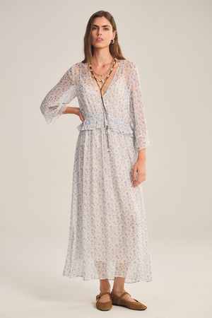 A woman in the AERIN DRESS by Velvet by Graham & Spencer—a romantic sheer maxi with long sleeves and a V-neck—pairs it with layered necklaces and brown flats, standing against a plain background.