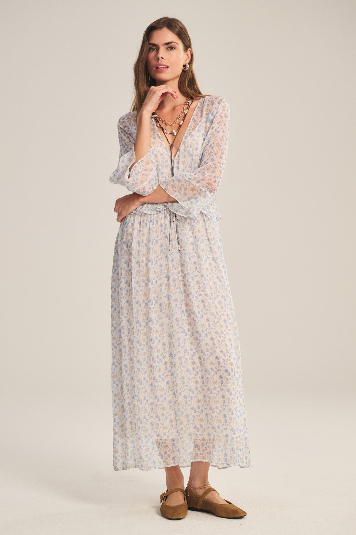 A woman stands against a plain background wearing the AERIN DRESS by Velvet by Graham & Spencer—a light, printed viscose maxi with long sleeves and a v-neck—styled with layered necklaces and tan flats for a romantic look.-40909523943617