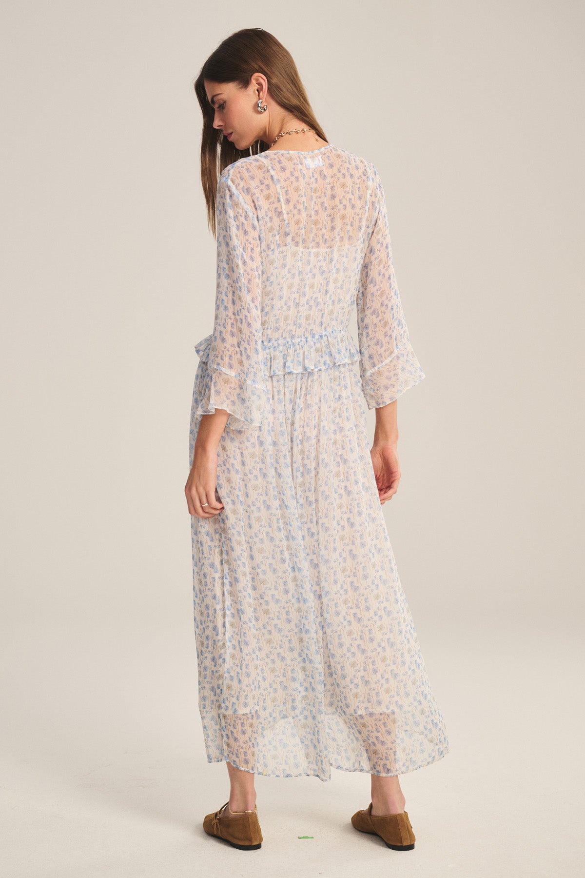 A woman stands facing away, wearing the AERIN DRESS by Velvet by Graham & Spencer— a sheer light blue floral maxi with three-quarter sleeves — and brown slip-on shoes, set against a neutral background.
