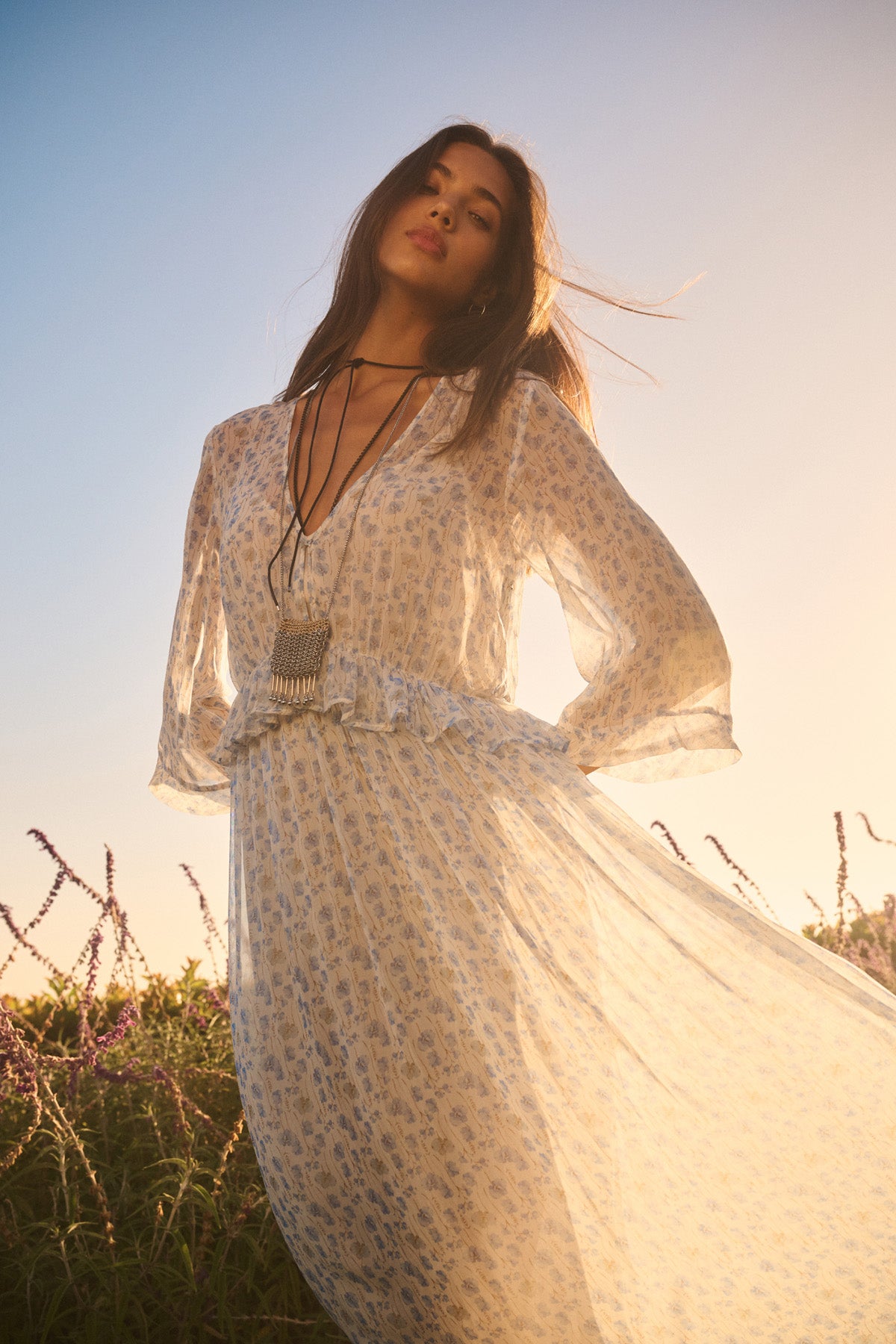 A woman stands outdoors at sunset in the AERIN DRESS by Velvet by Graham & Spencer, a sheer floral maxi with her hair down and layered necklaces—making a true romantic statement.-40909524140225