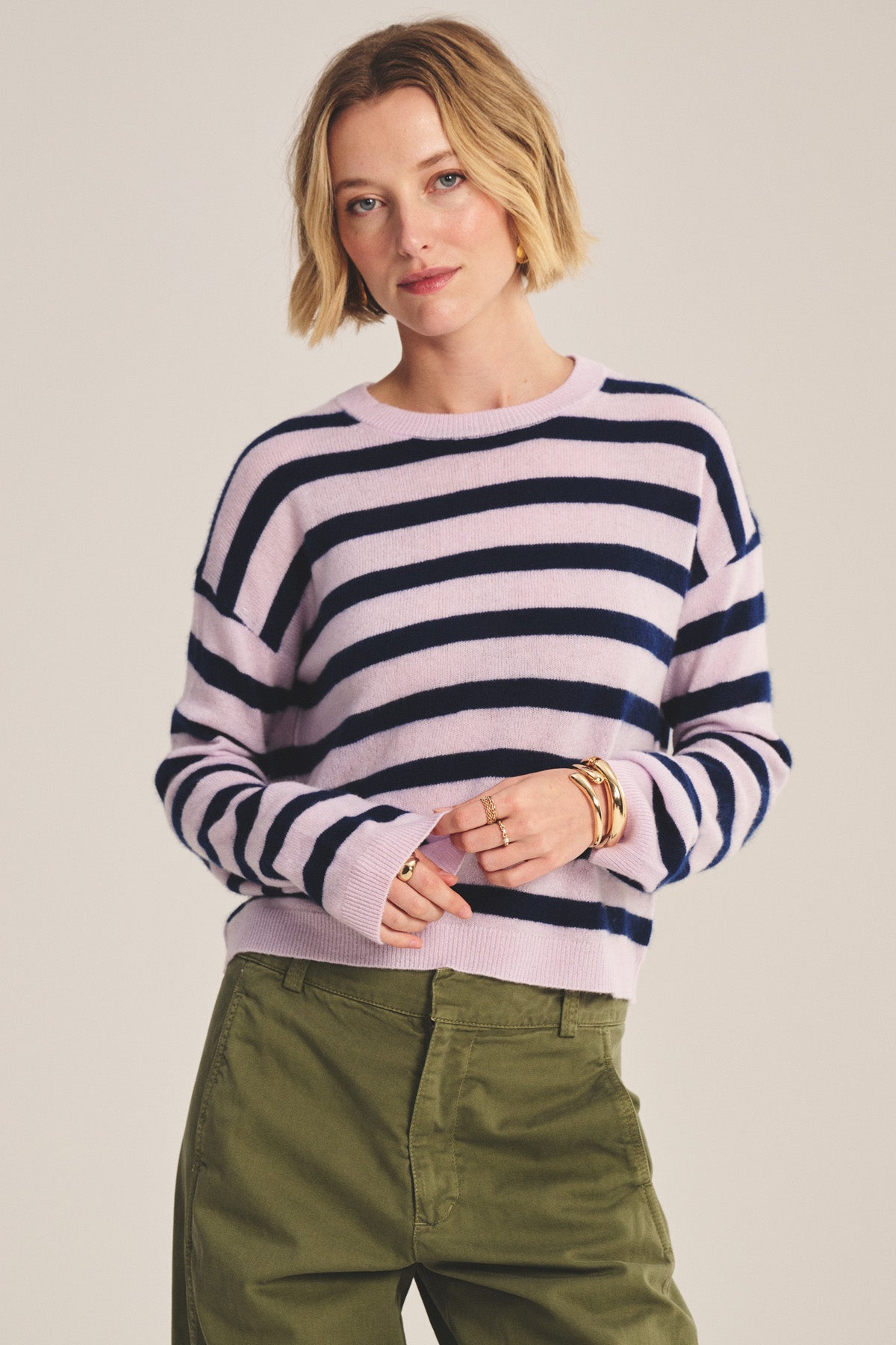 A woman with short blonde hair wears the Velvet by Graham & Spencer AGNESE Cashmere Sweater in light purple and navy stripes, paired with olive green pants—a versatile piece that provides lightweight warmth against a plain backdrop.-40808134672577