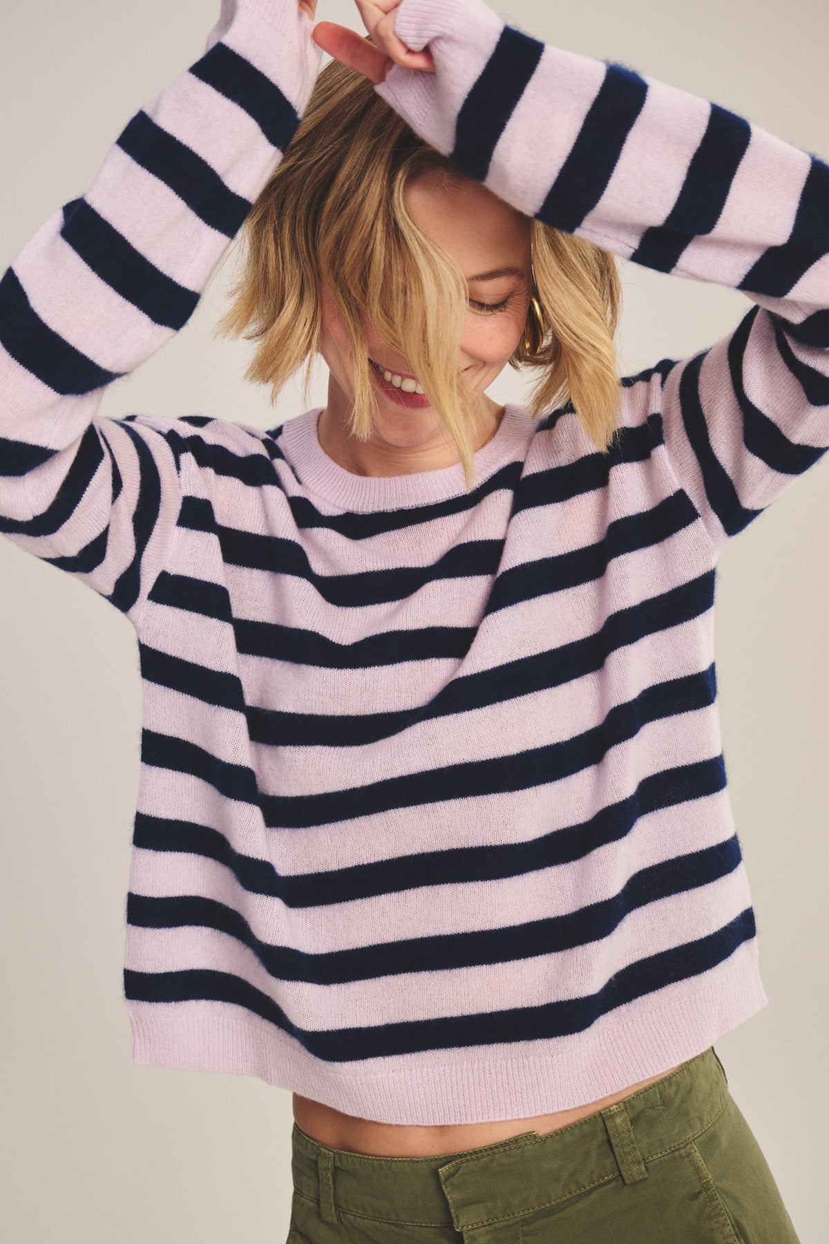 Wearing the AGNESE CASHMERE SWEATER by Velvet by Graham & Spencer, a person in pink and navy stripes with green pants raises their arms and smiles, hair partially covering their face.