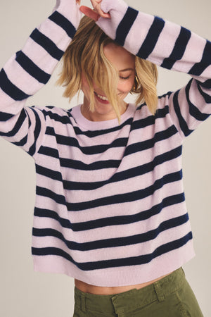 Wearing the AGNESE CASHMERE SWEATER by Velvet by Graham & Spencer, a person in pink and navy stripes with green pants raises their arms and smiles, hair partially covering their face.