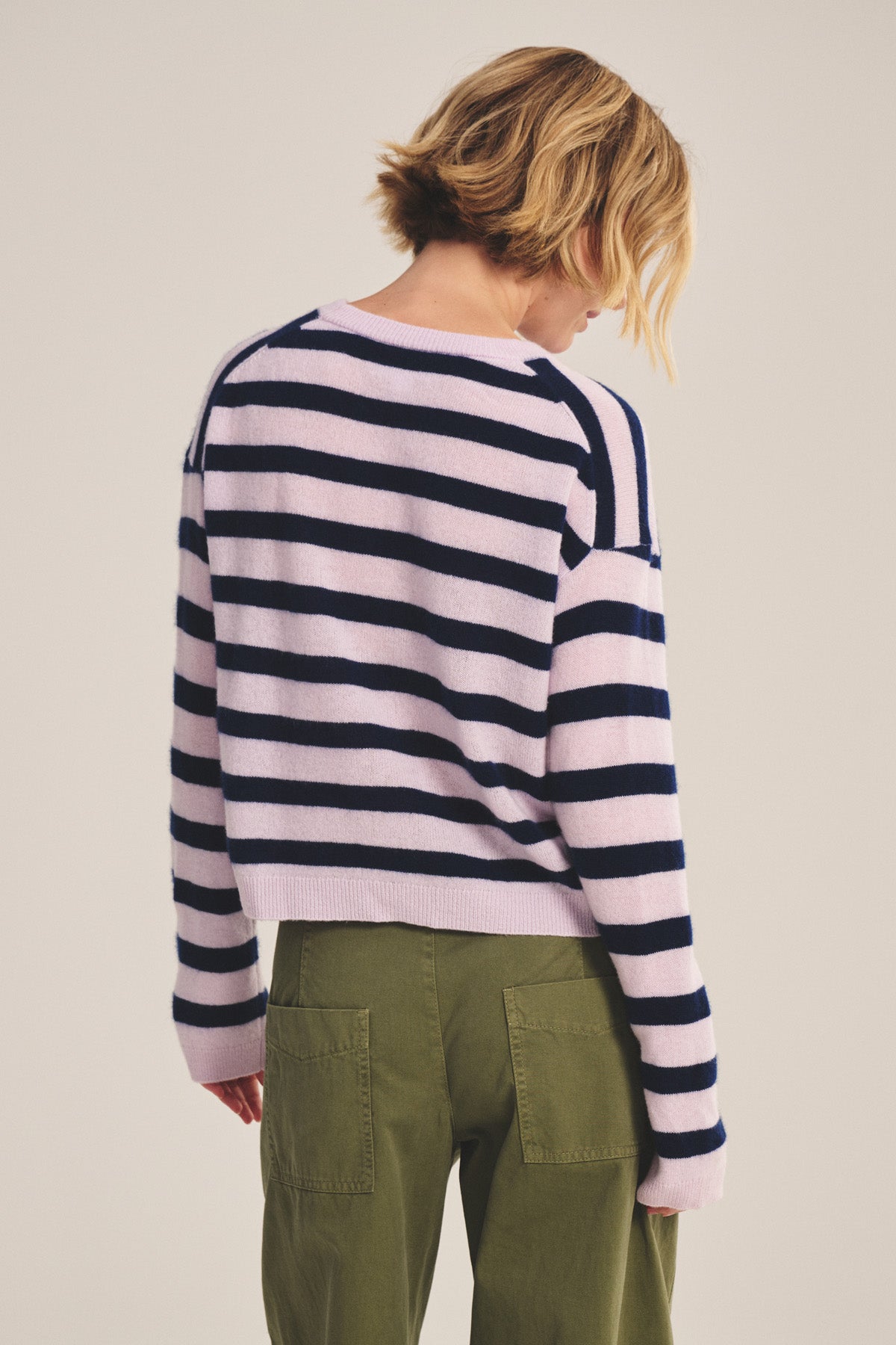 A person with short blonde hair is shown from the back, wearing Velvet by Graham & Spencer’s AGNESE CASHMERE SWEATER in light pink and navy stripes with olive green pants, against a plain background.-40808134836417