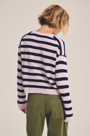 A person with short blonde hair is shown from the back, wearing Velvet by Graham & Spencer’s AGNESE CASHMERE SWEATER in light pink and navy stripes with olive green pants, against a plain background.