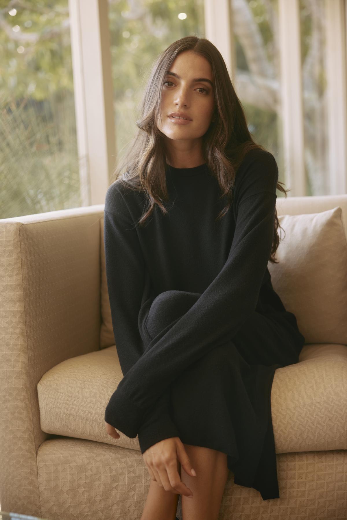   A person with long hair in the AIDA DRESS by Velvet by Graham & Spencer, featuring a detachable belt, sits on a beige sofa by a large window. 