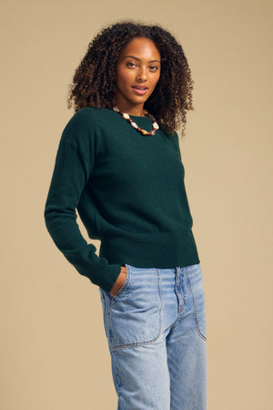 Wearing the Velvet by Graham & Spencer ALBA CASHMERE SWEATER in dark green, paired with light blue jeans and a chunky necklace, a person stands against a plain beige background with their hands in their pockets.