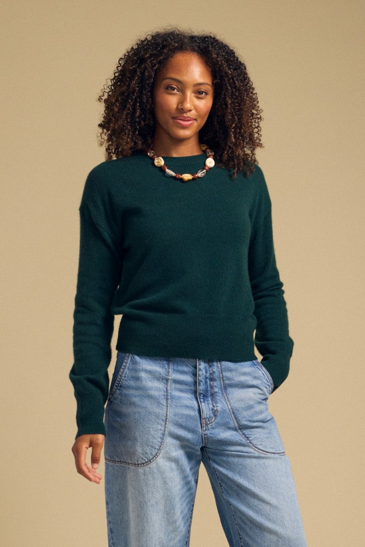 A woman with curly hair wears the ALBA CASHMERE SWEATER by Velvet by Graham & Spencer—a dark green, soft knit crewneck—paired with a beaded necklace and light blue jeans, standing against a plain beige background.