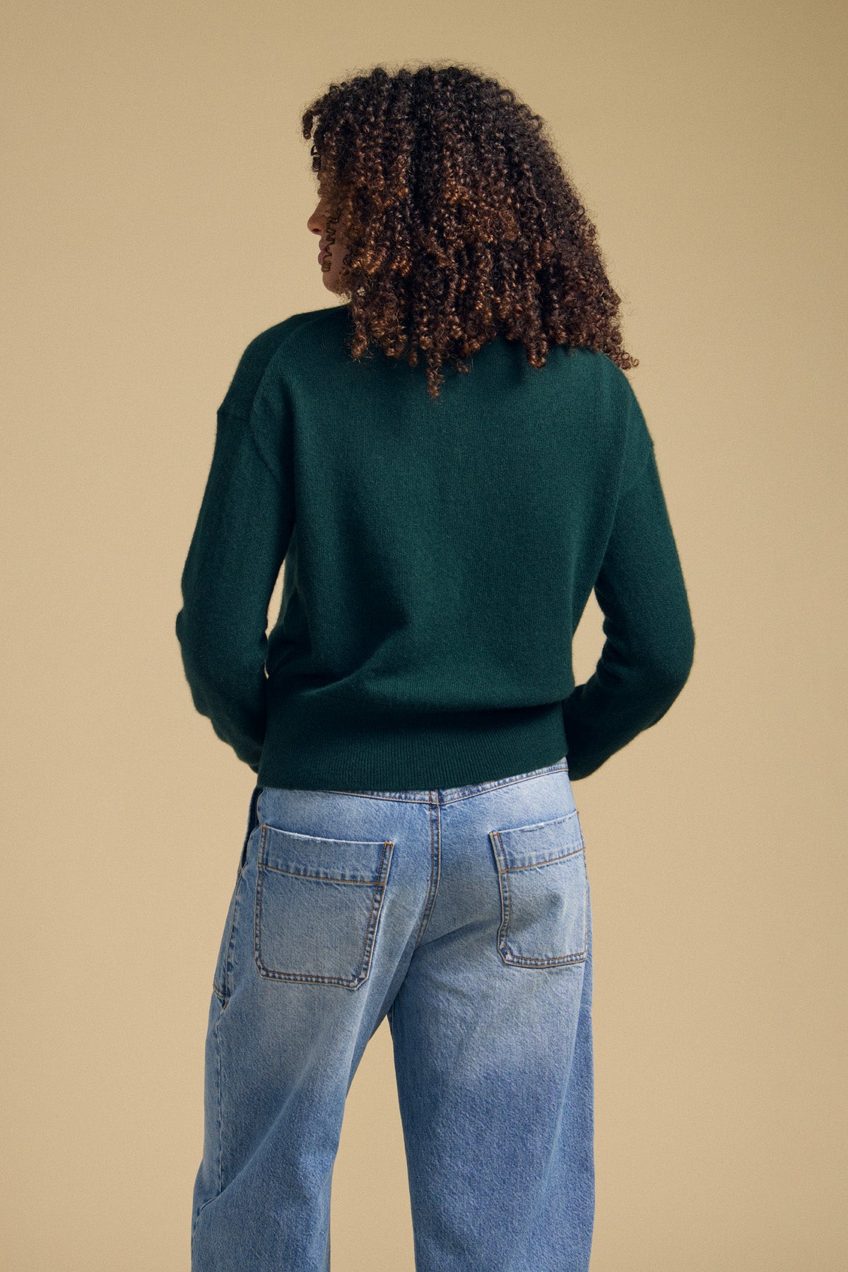 A person with curly hair is seen from behind, wearing the Velvet by Graham & Spencer ALBA CASHMERE SWEATER—a soft knit, dark green cashmere crewneck—paired with light blue jeans, set against a plain beige background.