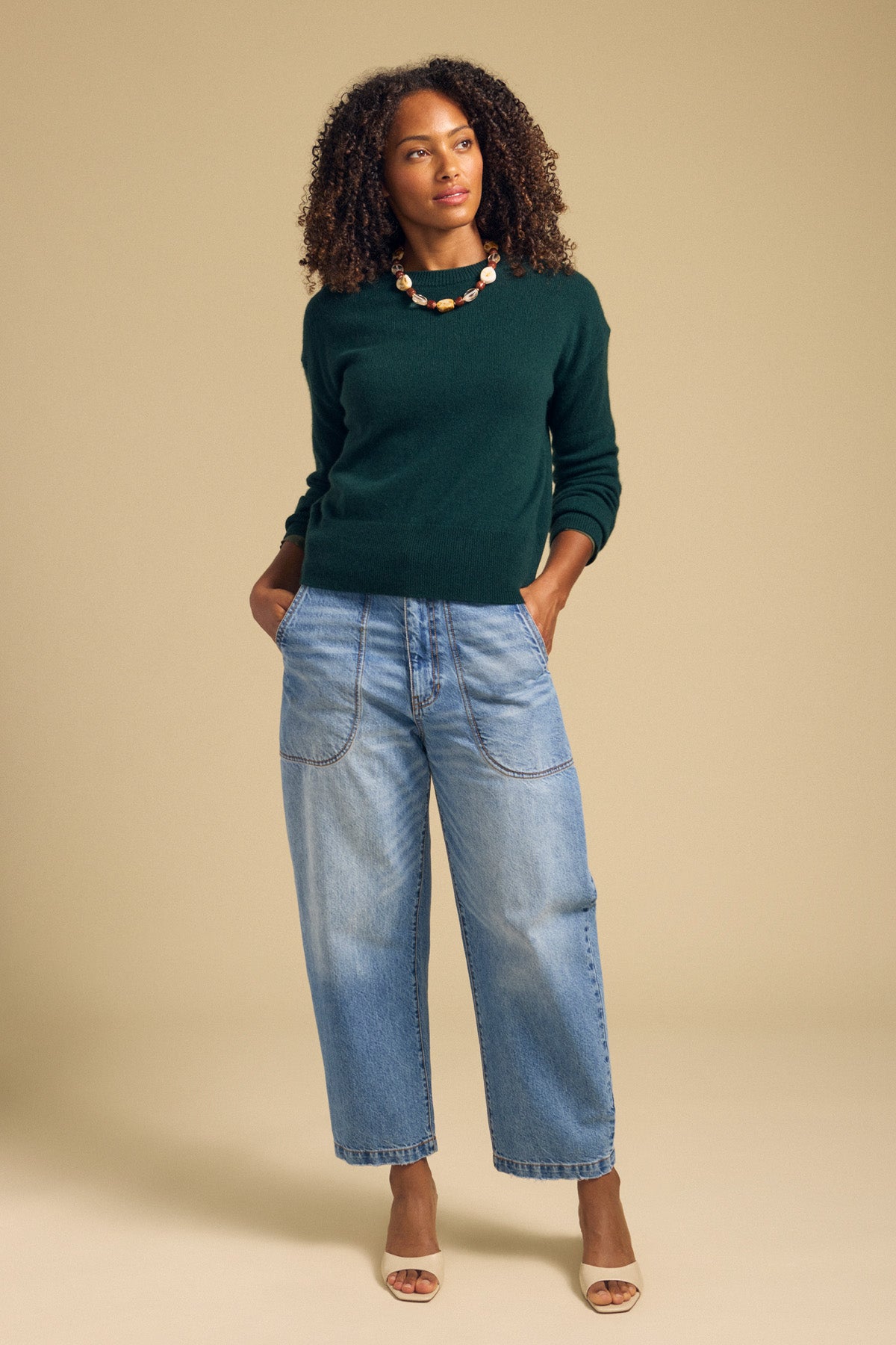 A woman stands against a beige background wearing the ALBA CASHMERE SWEATER by Velvet by Graham & Spencer in dark green, styled with light blue wide-leg jeans, a statement necklace, and beige heeled sandals, her hands tucked into her pockets.