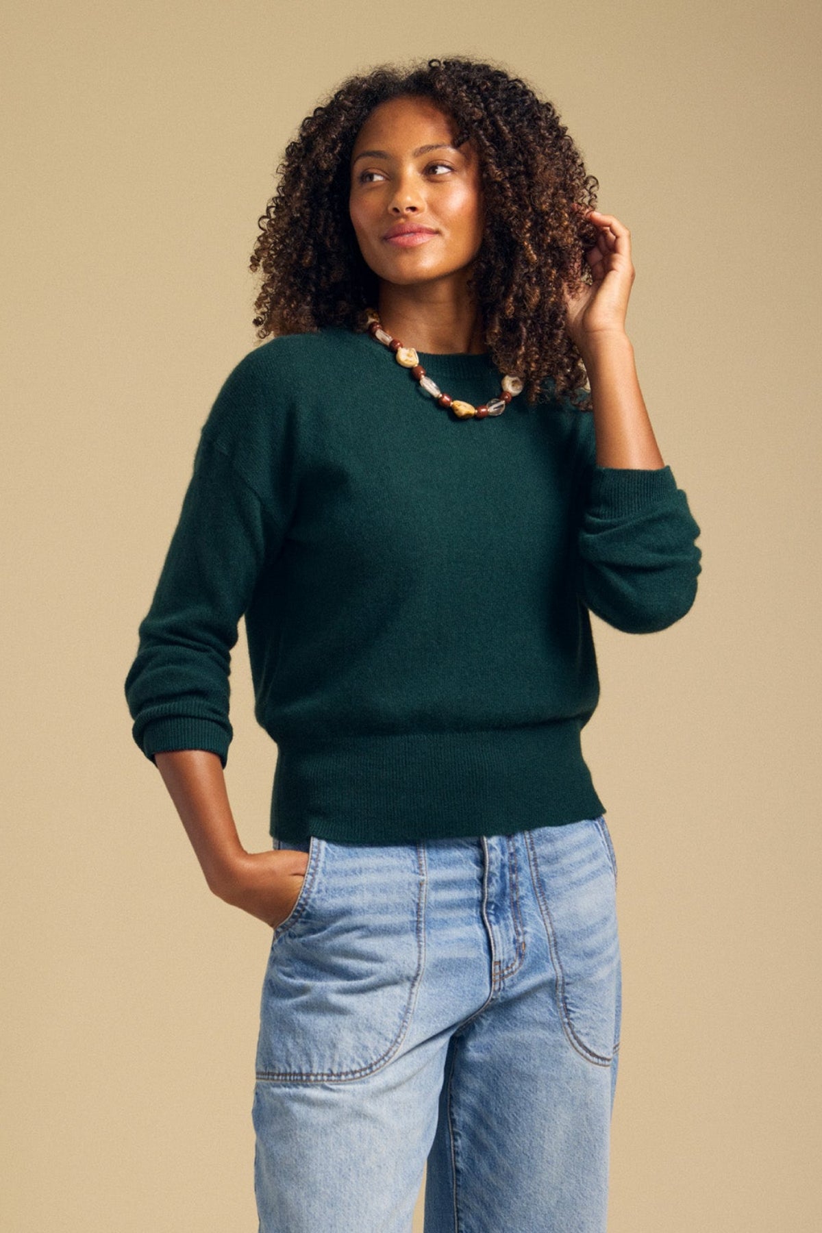 A woman with curly hair wears the Velvet by Graham & Spencer ALBA Cashmere Sweater in dark green, featuring a classic crew neckline, paired with light blue jeans and a chunky beaded necklace, standing against a plain beige background.