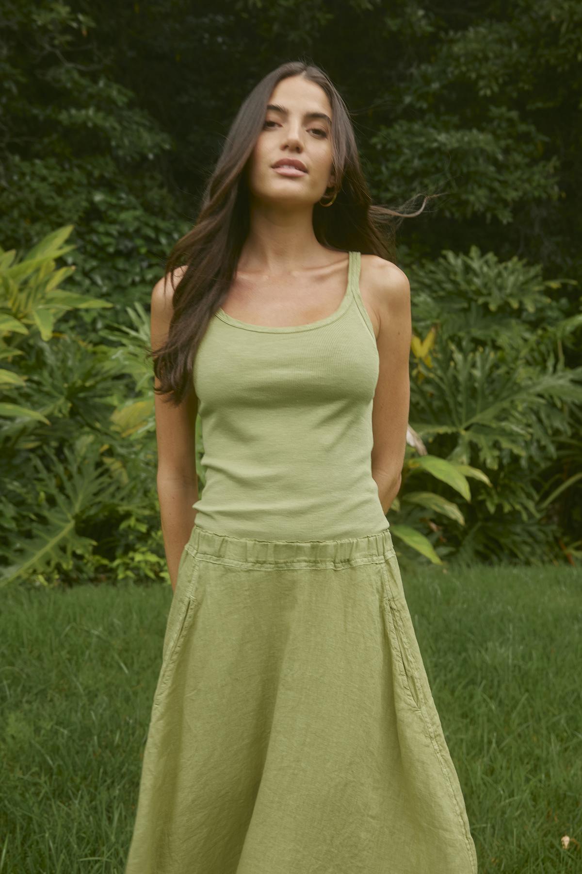   A woman stands in a lush outdoor setting wearing a light green tank top and an A-line skirt with an elastic waistband, the FAE LINEN SKIRT by Velvet by Graham & Spencer, both made from spring-weight linen. 