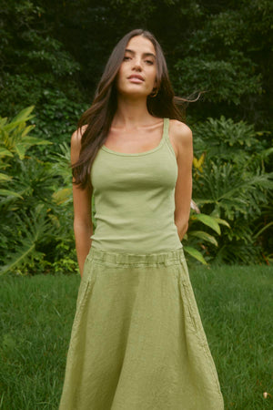 A woman with long dark hair stands outdoors wearing a green ALIZA RIBBED COTTON LAYERING TANK by Velvet by Graham & Spencer and a matching skirt. The background features lush green foliage.