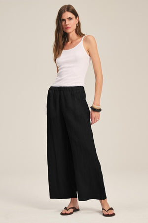 A woman stands with one hand on her hip against a neutral background, wearing a white tank top, sandals, and the effortless LOLA LINEN PANT by Velvet by Graham & Spencer—a versatile wide leg style for everyday wear.