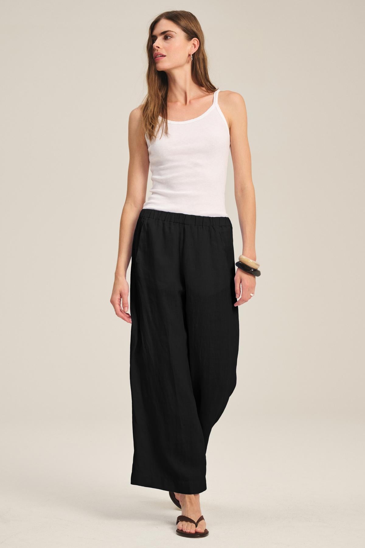 A woman stands against a plain background, wearing a white tank top, black sandals, bracelets, and the LOLA LINEN PANT by Velvet by Graham & Spencer, looking slightly to the side.