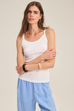 A woman stands against a plain background wearing the ALIZA RIBBED COTTON LAYERING TANK by Velvet by Graham & Spencer, paired with light blue pants and bracelets on her left wrist.