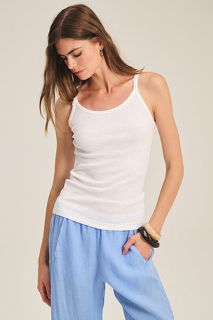 A woman wearing the ALIZA RIBBED COTTON LAYERING TANK by Velvet by Graham & Spencer with light blue pants stands against a plain background, looking down with one hand resting by her side.