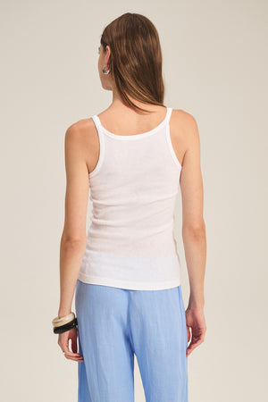 A person with long hair stands with their back to the camera, wearing the ALIZA RIBBED COTTON LAYERING TANK by Velvet by Graham & Spencer and light blue pants against a plain background.