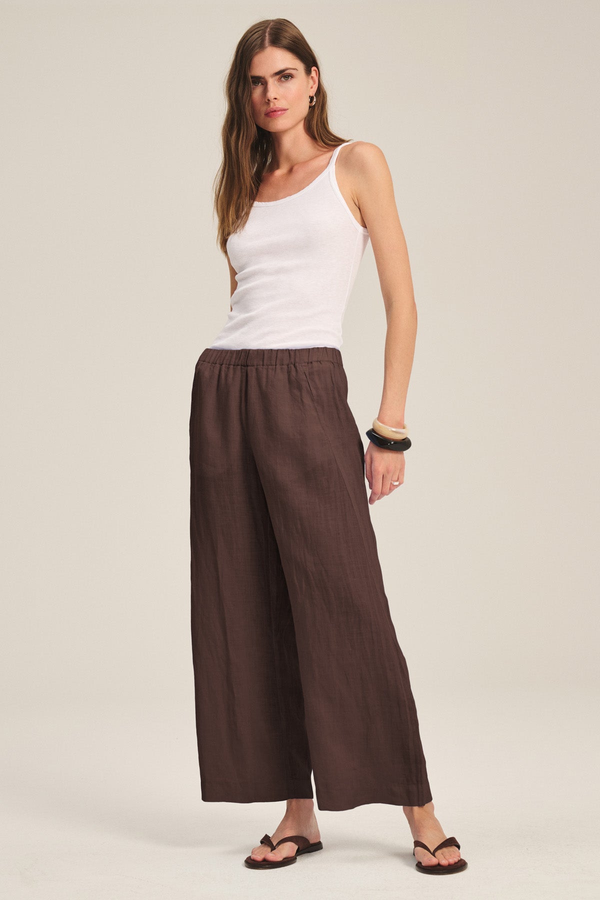 A woman wears the LOLA LINEN PANT by Velvet by Graham & Spencer—loose, lightweight linen pants with an elastic waist—paired with a white sleeveless top, black sandals, and bracelets against a plain background.