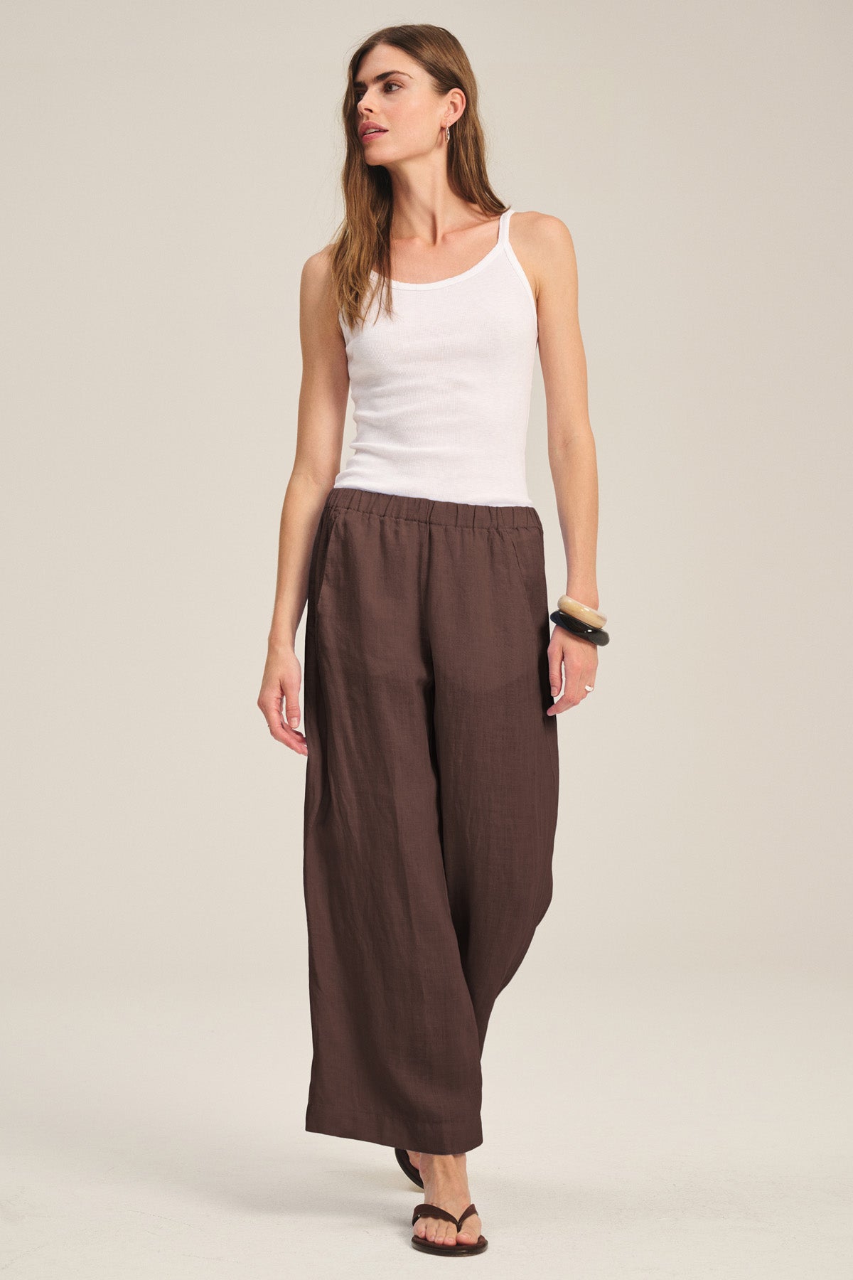 A woman stands against a plain light background wearing a white tank top, sandals, and the LOLA LINEN PANT in loose brown lightweight linen by Velvet by Graham & Spencer.