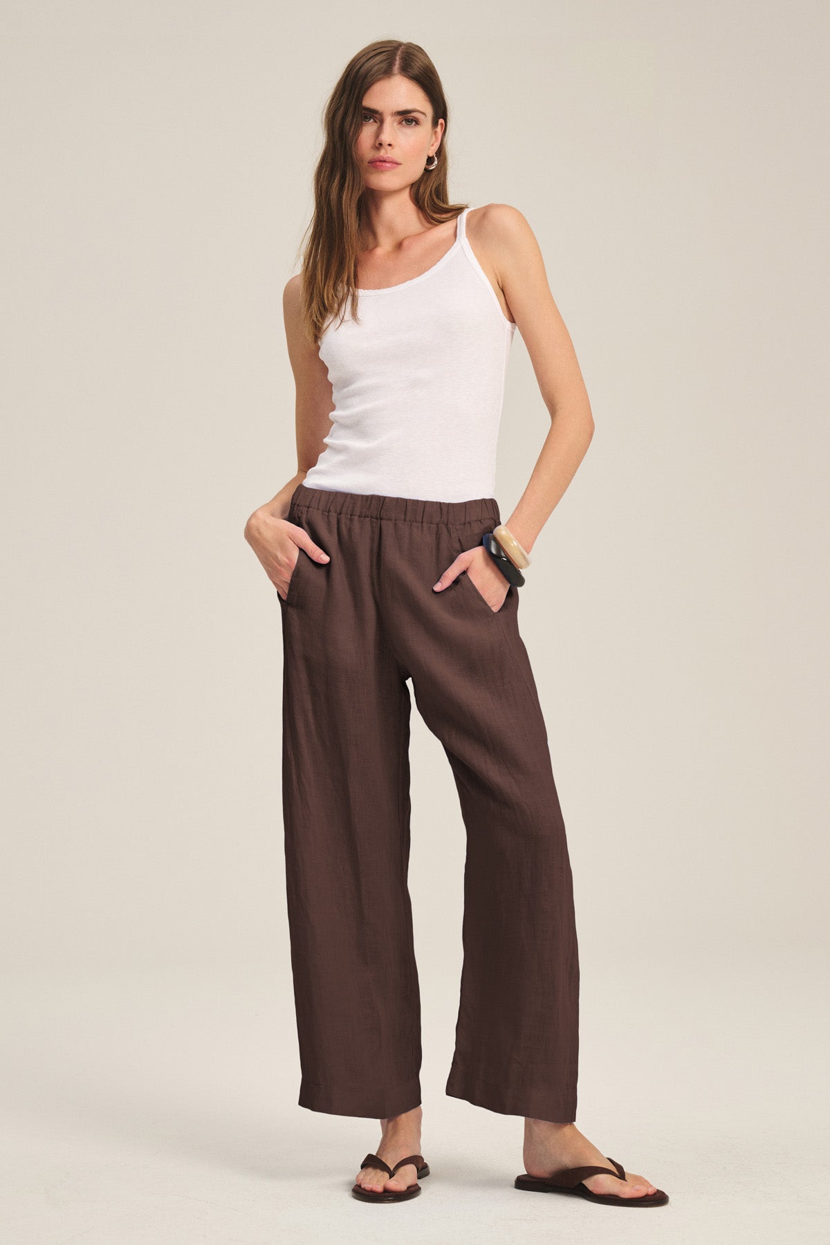 A woman stands against a neutral background wearing the LOLA LINEN PANT by Velvet by Graham & Spencer, styled with a white tank top, black sandals, and bracelets, her hands in the pant's pockets.-40673323253953