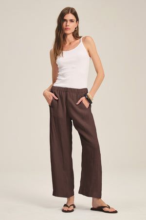 A woman stands against a neutral background wearing the LOLA LINEN PANT by Velvet by Graham & Spencer, styled with a white tank top, black sandals, and bracelets, her hands in the pant's pockets.