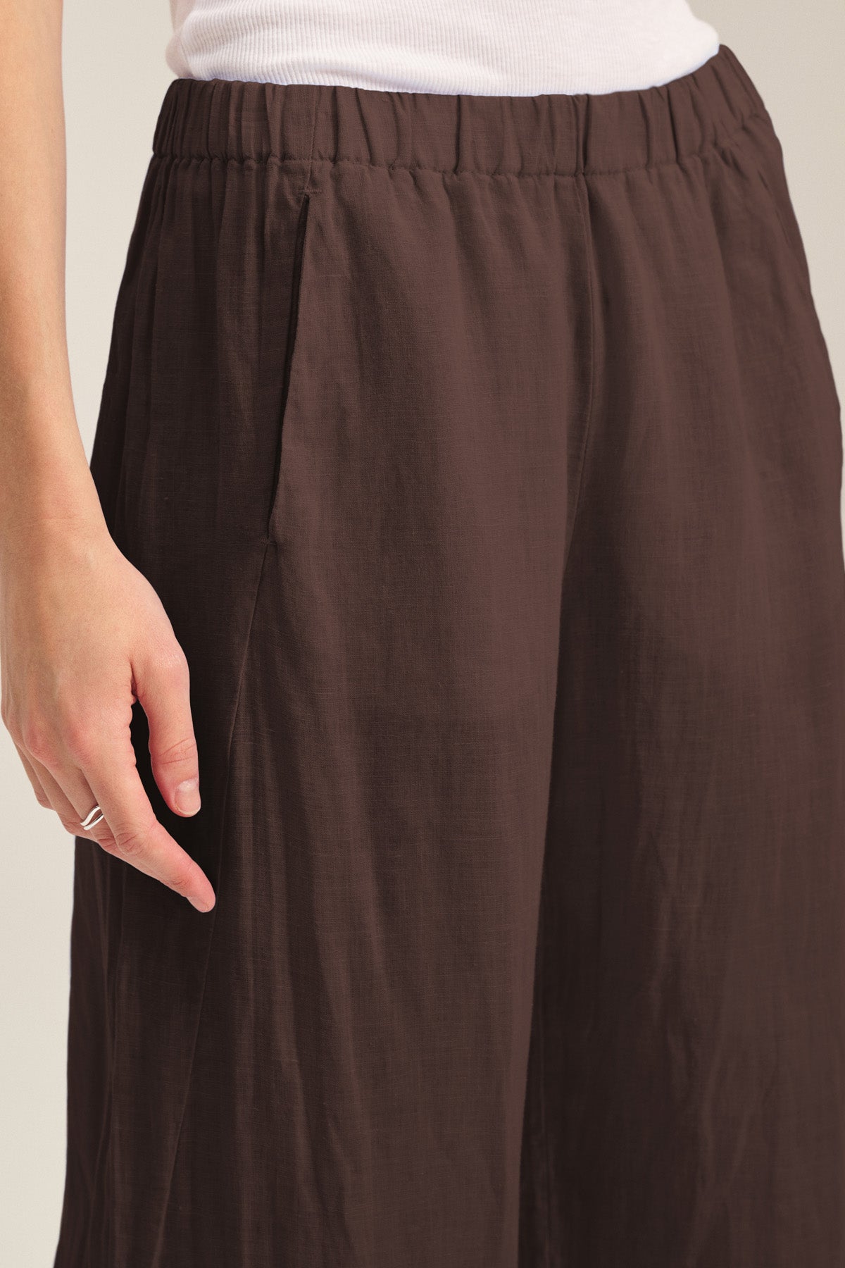 A person wearing the LOLA LINEN PANT by Velvet by Graham & Spencer—a brown, lightweight linen pant with an elastic waist—stands with their left hand resting at their side, paired with a white top.