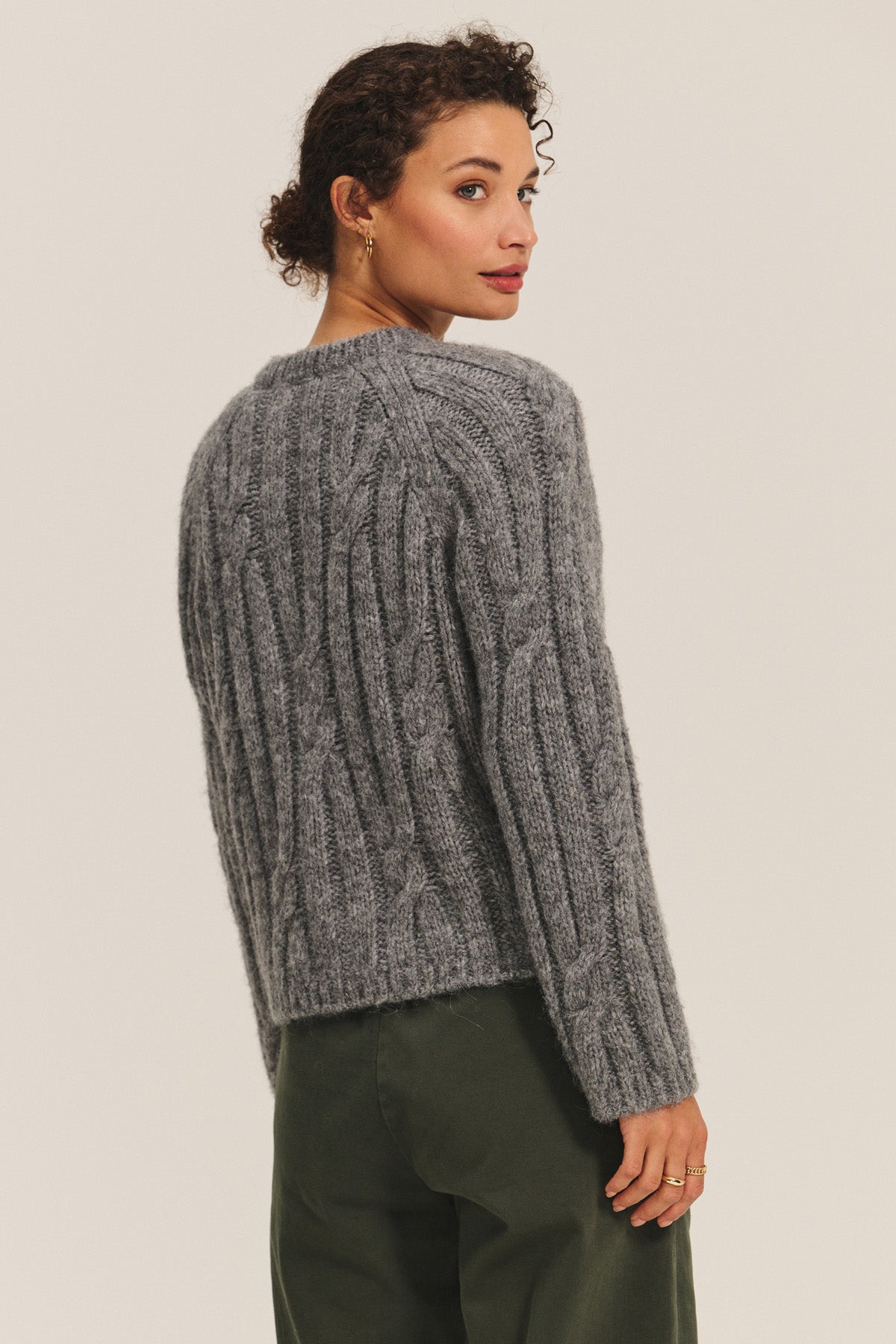 A woman with short curly hair wears the ALLISON CARDIGAN by Velvet by Graham & Spencer in chunky gray cable-knit, paired with dark green pants—an effortlessly stylish, cozy layering piece—while standing against a plain background.
