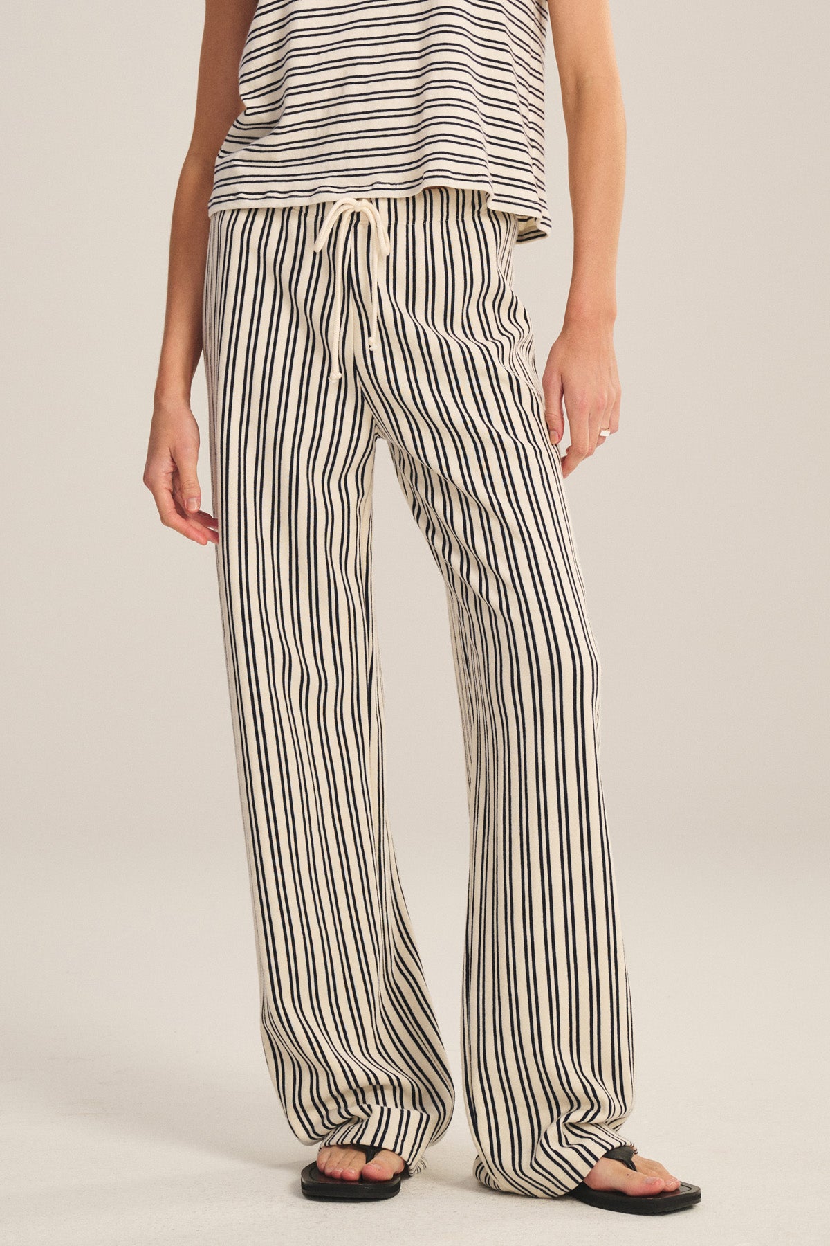 A person stands against a plain background wearing the AMELISE SWEATPANT by Velvet by Graham & Spencer, featuring black-and-white vintage stripes, a drawstring elastic waist, and a relaxed straight-leg fit, paired with a matching sleeveless top and flip-flops.