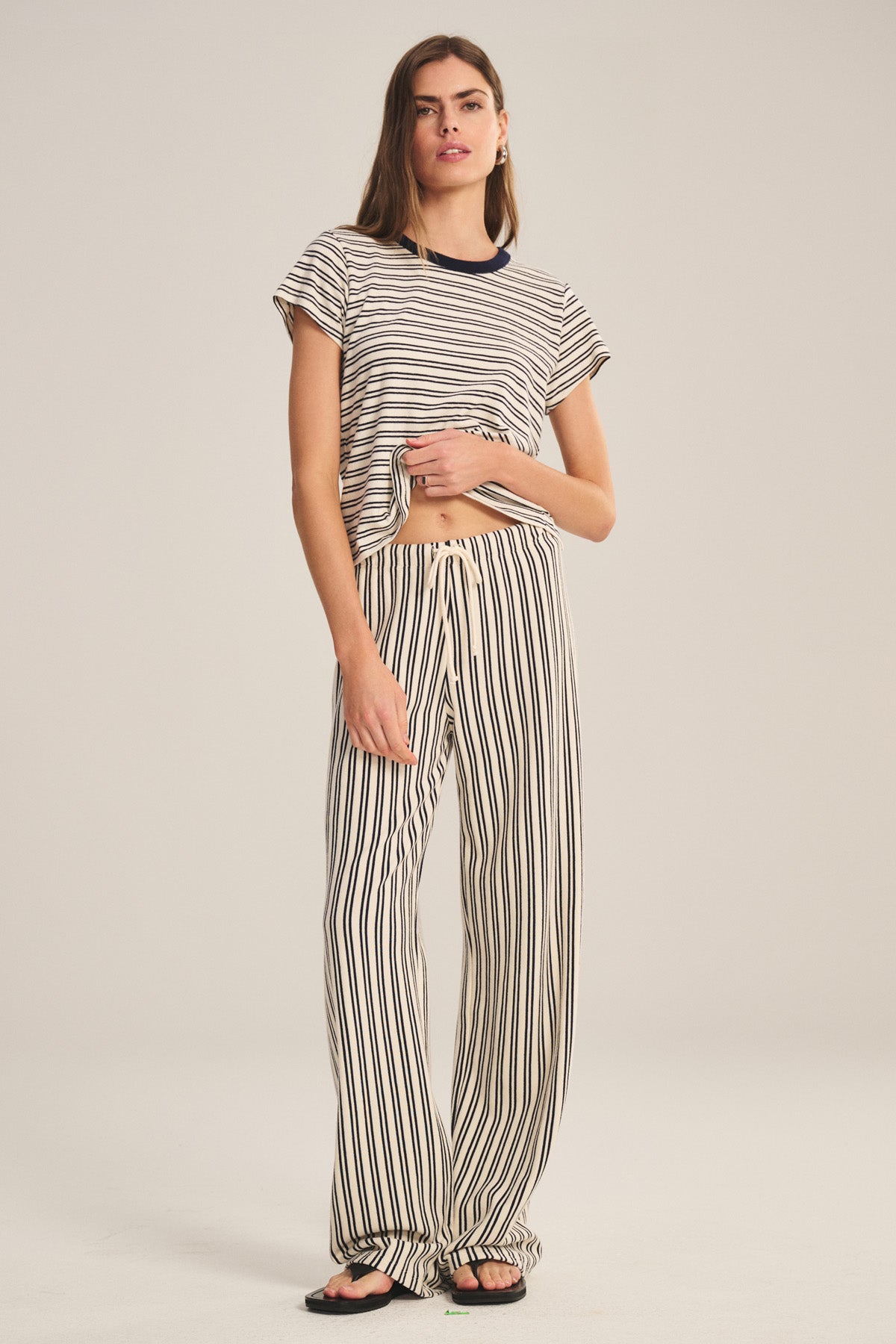 A woman stands against a plain background wearing the AMELISE SWEATPANT by Velvet by Graham & Spencer, featuring a relaxed straight-leg fit, drawstring elastic waist, and paired with a short-sleeve striped tee and black sandals.-40909622116545
