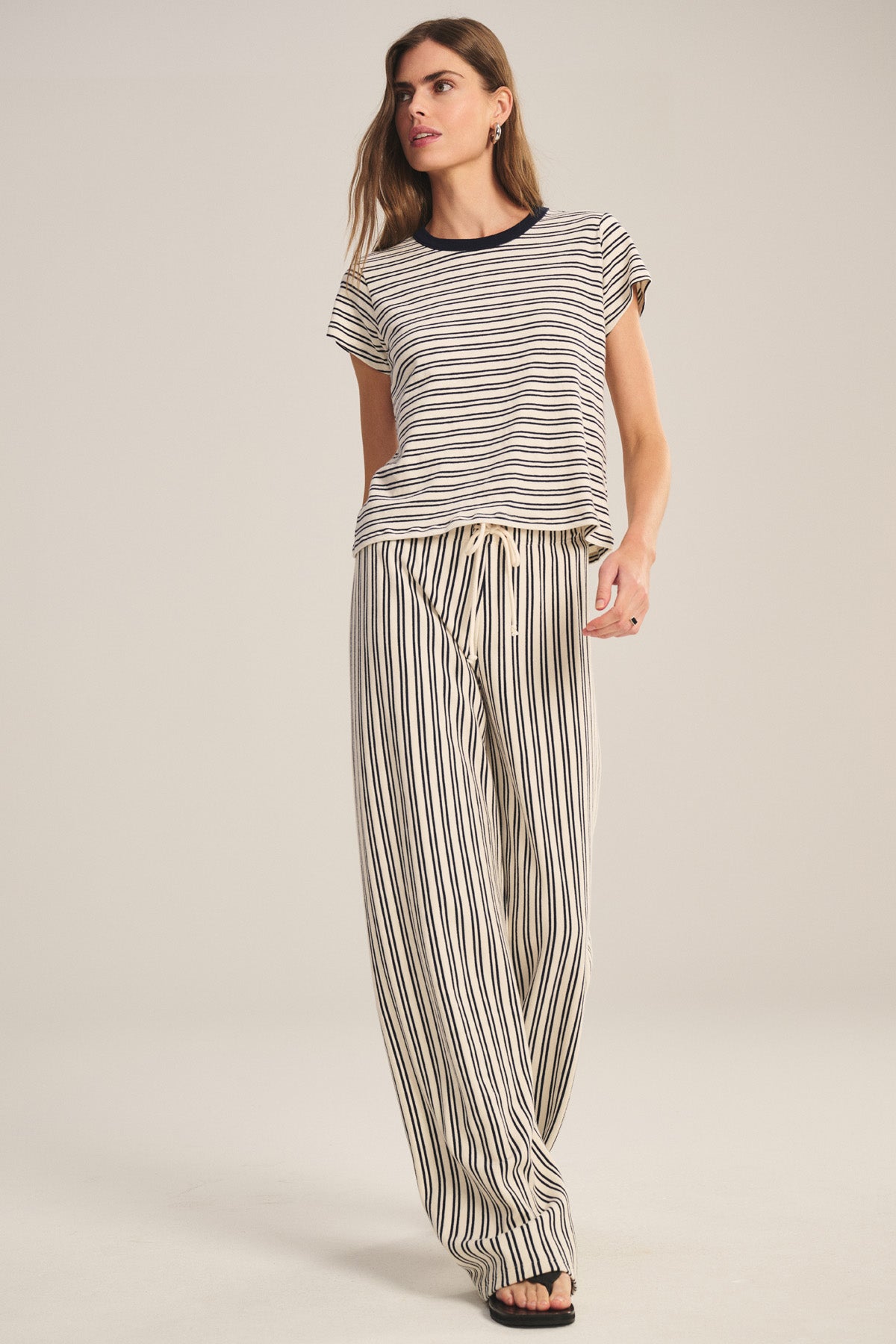A woman stands against a plain background wearing the Velvet by Graham & Spencer AMELISE SWEATPANT, a vintage stripe terry pant with drawstring elastic waist and relaxed straight-leg fit, paired with a matching short-sleeved striped top and black sandals.-40909622149313