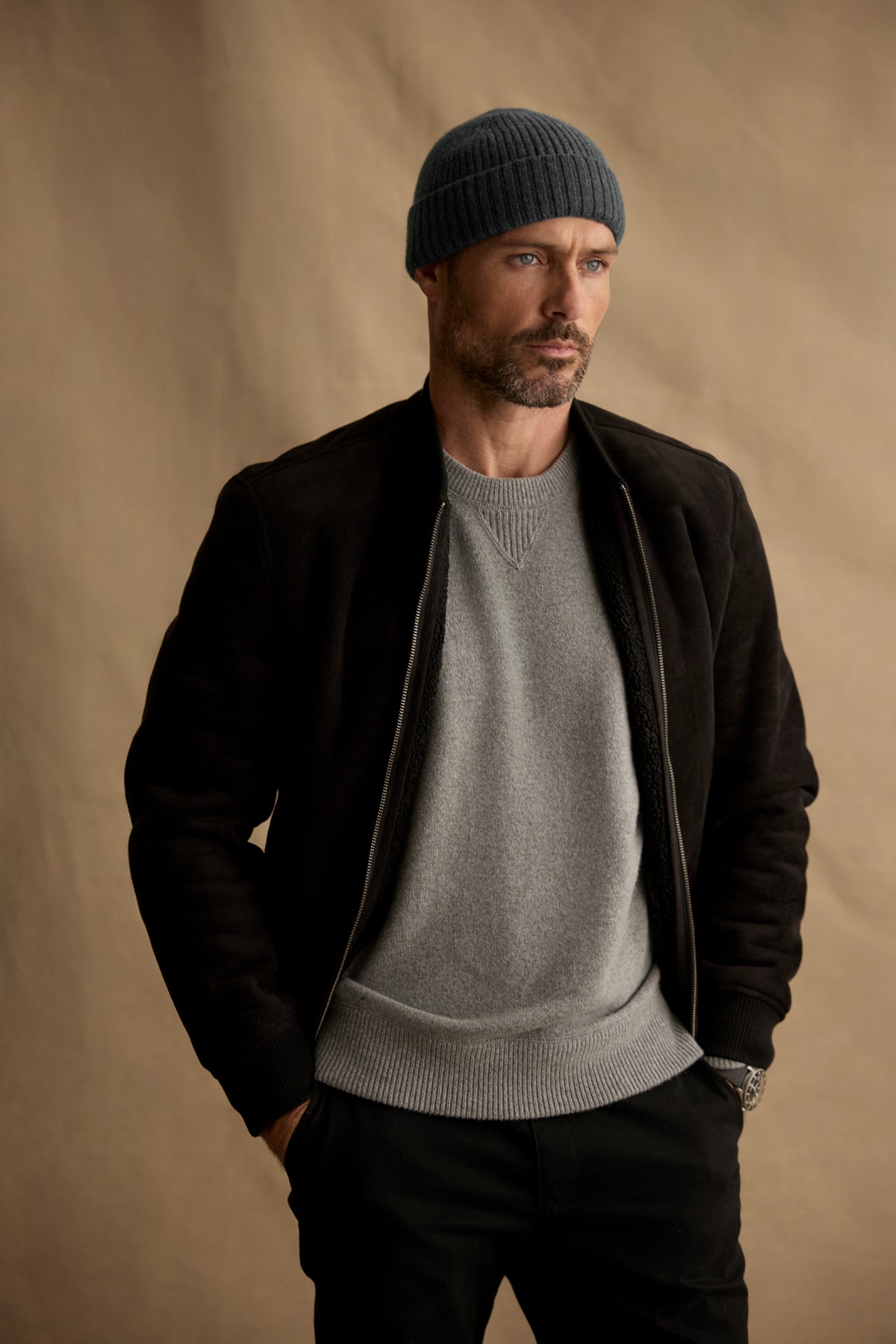 Man wears the AMIAS BOMBER JACKET by Velvet by Graham & Spencer—faux suede with sherpa lining—paired with a gray knit beanie and sweater, hands in pockets against a beige background for an understated men’s fashion look.-40054203580609
