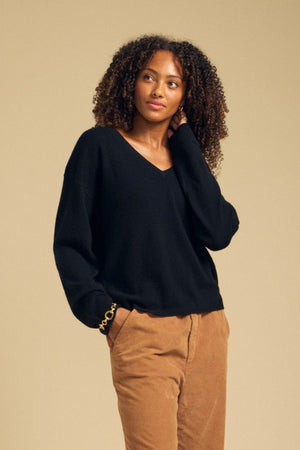 A person with curly hair wears the AMIKA CASHMERE SWEATER by Velvet by Graham & Spencer—featuring a deep V neckline in black—paired with tan pants, standing against a plain beige background with one hand touching their hair.