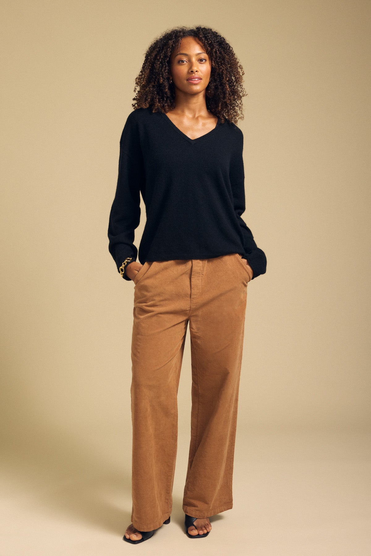 A woman stands against a beige background wearing the AMIKA CASHMERE SWEATER by Velvet by Graham & Spencer—a black cashmere sweater featuring a deep V neckline—paired with tan wide-leg pants with a cropped waist and black open-toe shoes, her hands resting in her pockets.