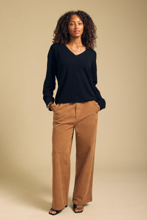A woman stands against a beige background wearing the AMIKA CASHMERE SWEATER by Velvet by Graham & Spencer—a black cashmere sweater featuring a deep V neckline—paired with tan wide-leg pants with a cropped waist and black open-toe shoes, her hands resting in her pockets.