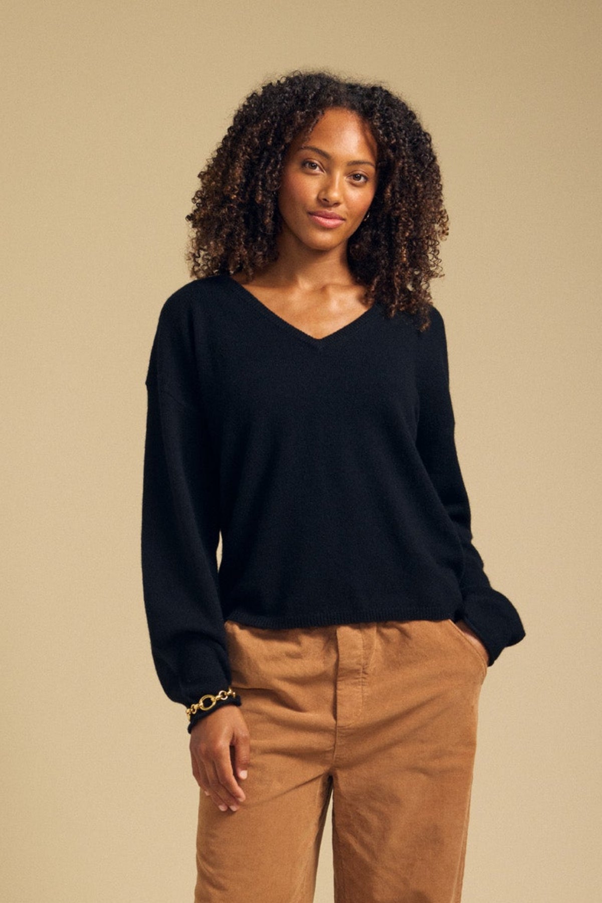 A woman with curly hair wears the Velvet by Graham & Spencer AMIKA Cashmere Sweater in black, featuring a deep V neckline, paired with tan pants. She stands against a plain beige background with a neutral expression.