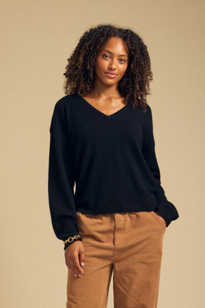 A woman with curly hair wears the Velvet by Graham & Spencer AMIKA Cashmere Sweater in black, featuring a deep V neckline, paired with tan pants. She stands against a plain beige background with a neutral expression.