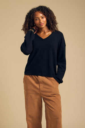 A woman with curly hair stands against a beige background, wearing the AMIKA CASHMERE SWEATER by Velvet by Graham & Spencer—a black cashmere sweater featuring a deep V neckline—paired with brown pants and one hand in her pocket.