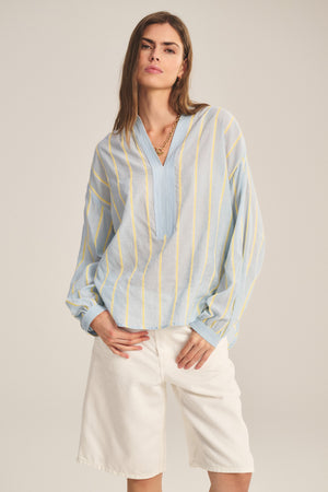 A woman wears the ANALYN TOP by Velvet by Graham & Spencer—a light blue yarn-dyed stripe cotton, long-sleeve blouse with yellow accents and a V-neckline—paired with white knee-length shorts, standing against a plain background.