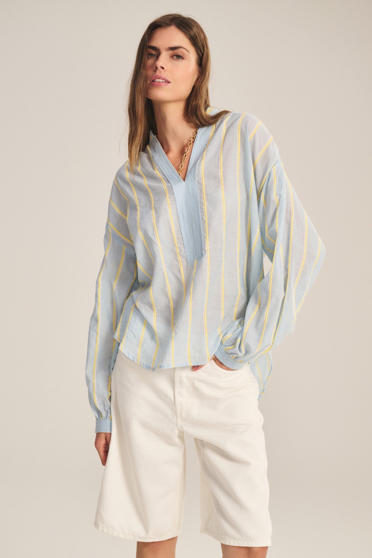 A woman wears the Velvet by Graham & Spencer ANALYN TOP, a light blue relaxed fit with yellow yarn-dyed stripes, paired with white knee-length shorts against a plain background.-40959848939713