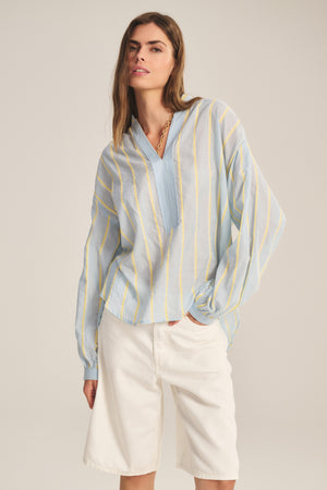 A woman wears the Velvet by Graham & Spencer ANALYN TOP, a light blue relaxed fit with yellow yarn-dyed stripes, paired with white knee-length shorts against a plain background.